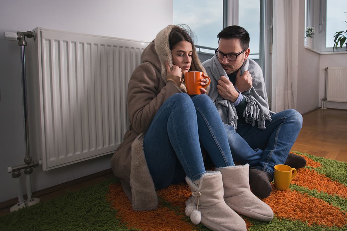 Ein Mann und eine Frau sitzen mit Tassen in der Hand in einer kalten Wohnung am Heizkörper und sind mit Decken bedeckt